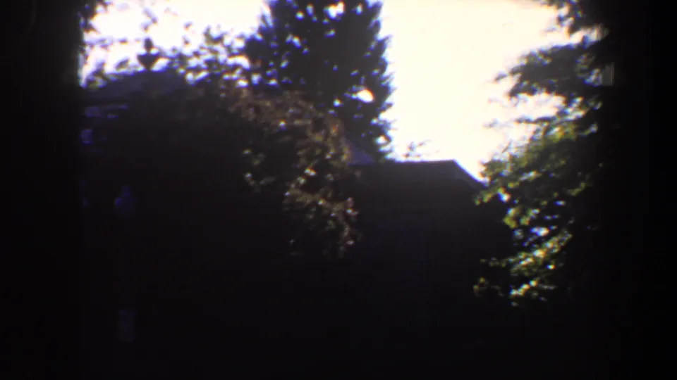 1964 8mm frame showing a dark house exterior behind trees, bushes, and a bright washed-out sky.