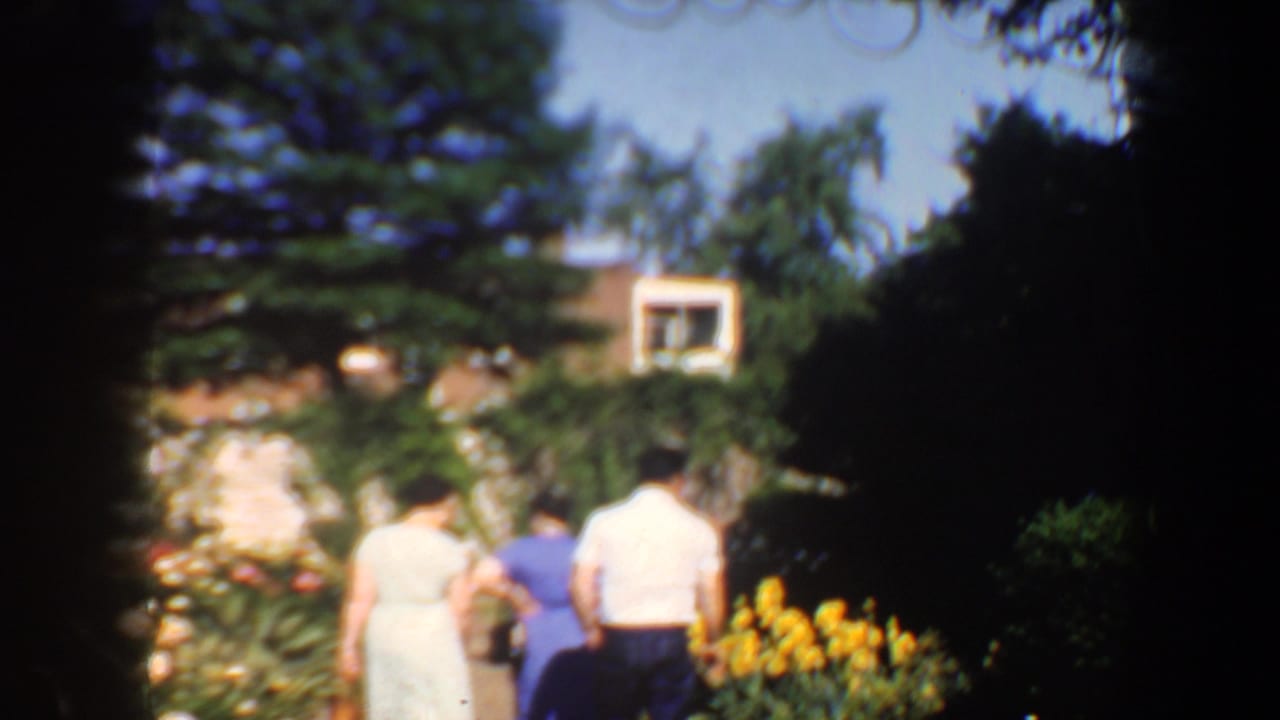 Three people walk away from the camera through a flower garden toward a house.