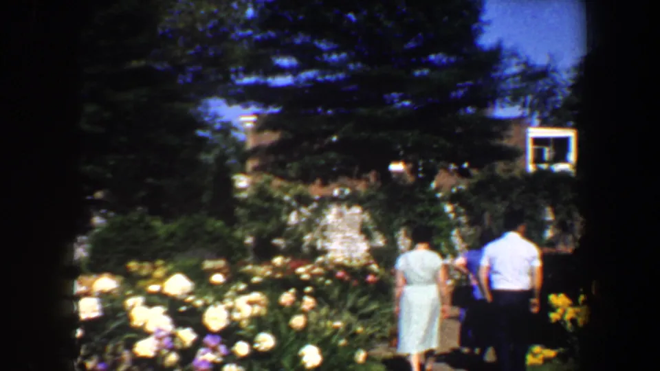 A rear-view family group moves along a landscaped garden path with abundant flowers in the foreground and trees overhead.