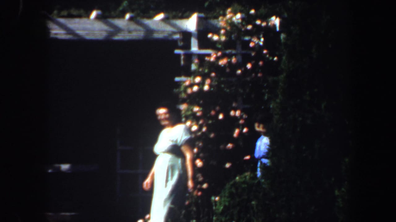 Woman in a pale dress walking near flowers and shaded garden structures.