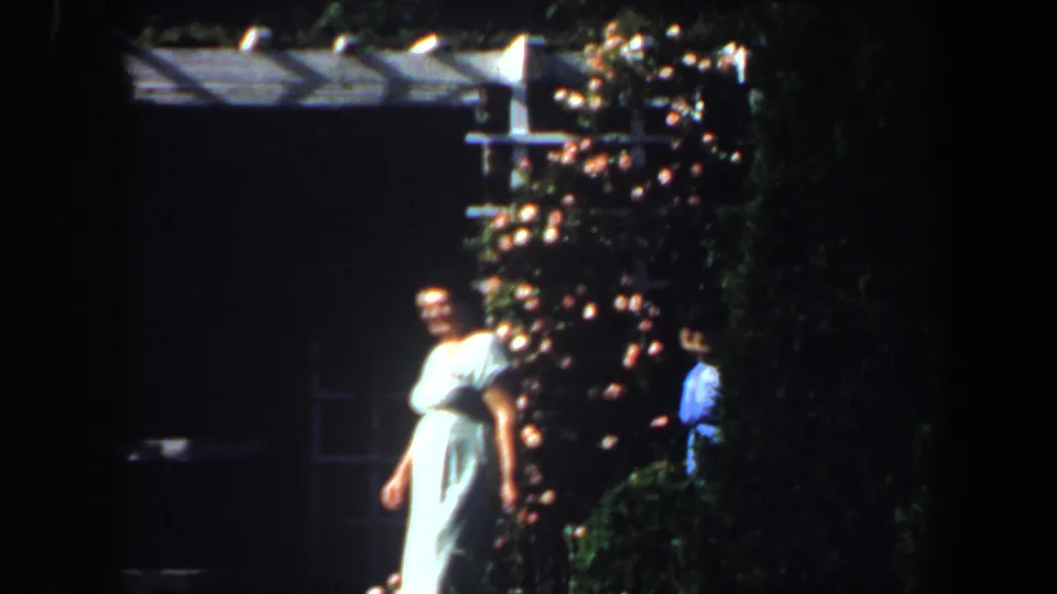1964 Saint Louis garden scene with a woman in a pale dress moving past flowering plants and deep shade.