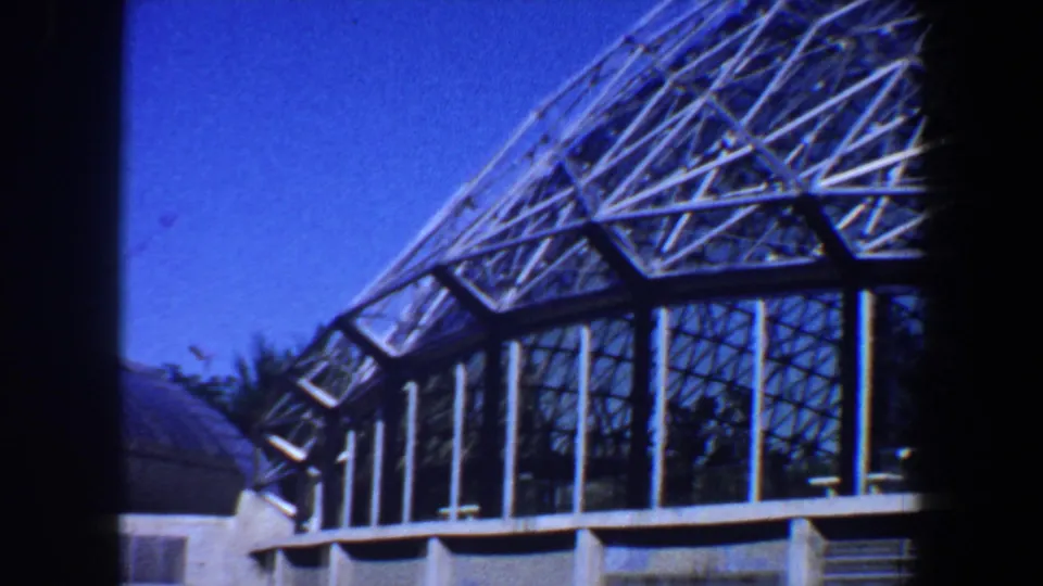 1964 Saint Louis frame looking across the angular glass facade, vertical supports, and nearby roof under clear daylight.