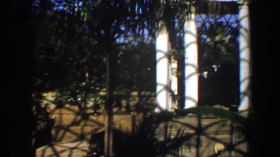 1964 Saint Louis home-movie frame showing the conservatory's white columns, plants, low barrier, and strong lattice shadows across the scene.