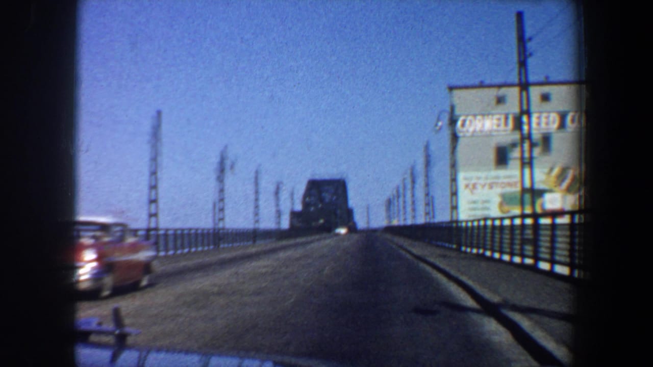 Forward car view on a bridge with a red car passing and a Cornell Seed Co. billboard at right.