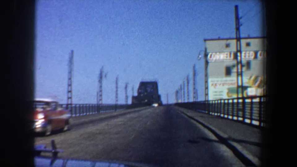 1964 Saint Louis bridge crossing seen from inside a car, with road railings, a red car, and Cornell Seed Co. signage visible.