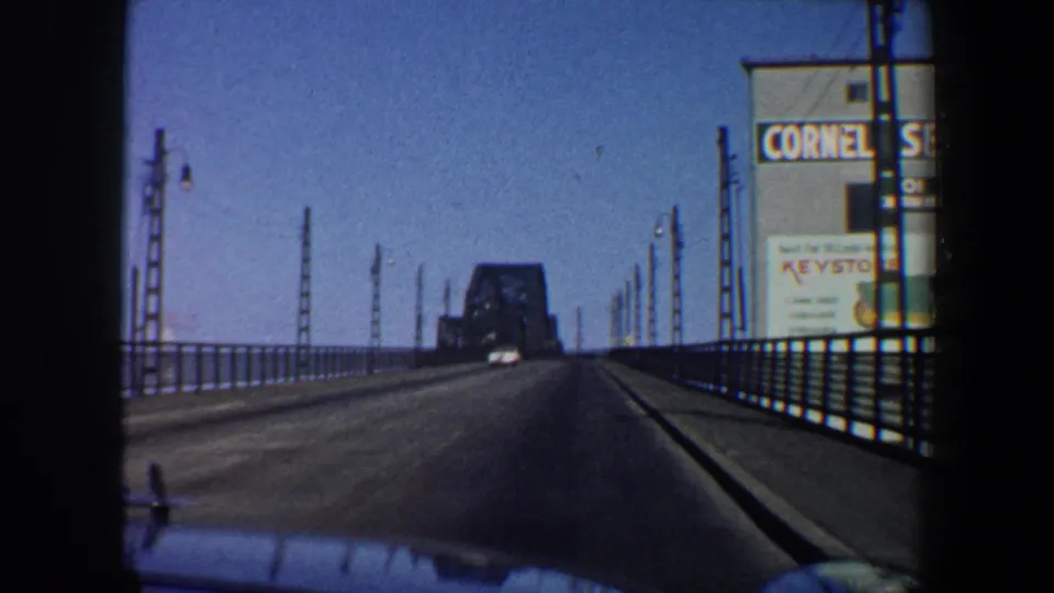 Late frame from the 1964 Saint Louis bridge drive, with the car hood, open roadway, railings, poles, and Cornell Seed Co. billboard.