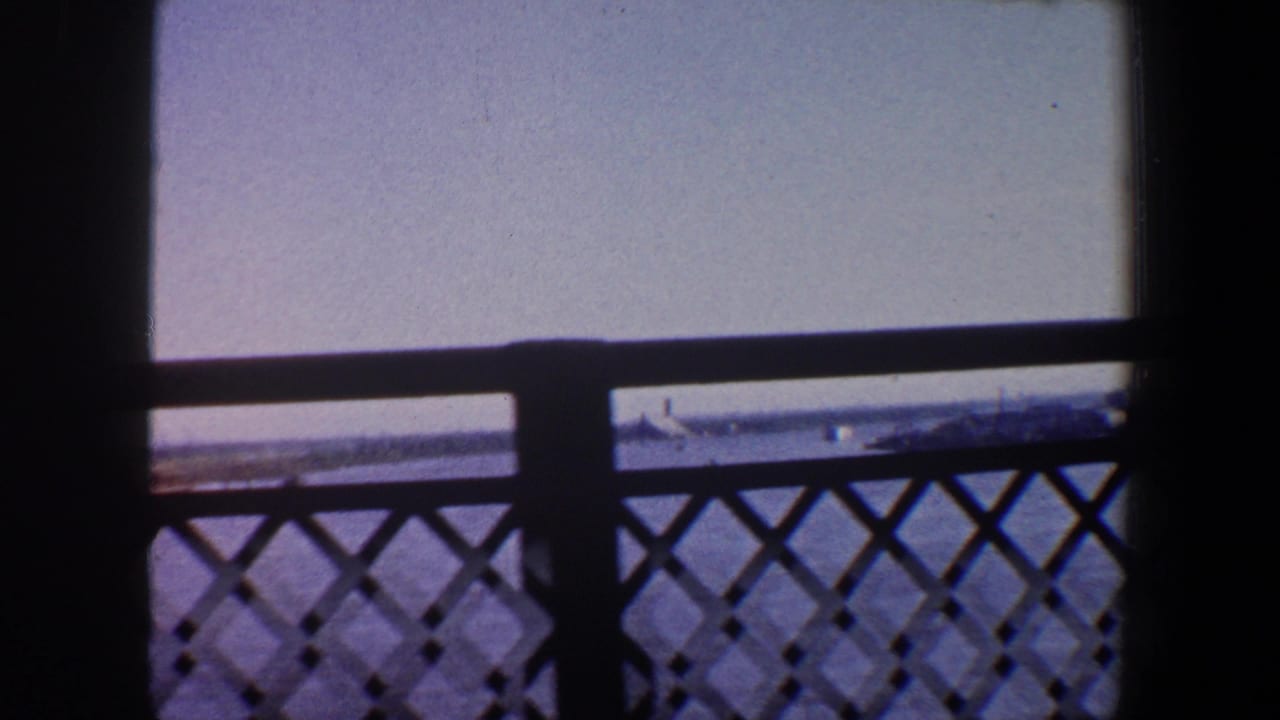 Bridge railing in silhouette with river, pale sky, and distant shoreline beyond.