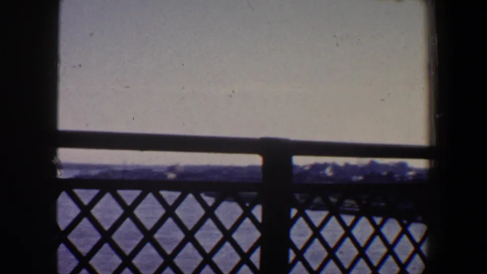 A 1964 8mm view from a Saint Louis bridge shows river water, a dark lattice railing, and a low shoreline under a washed pale sky.