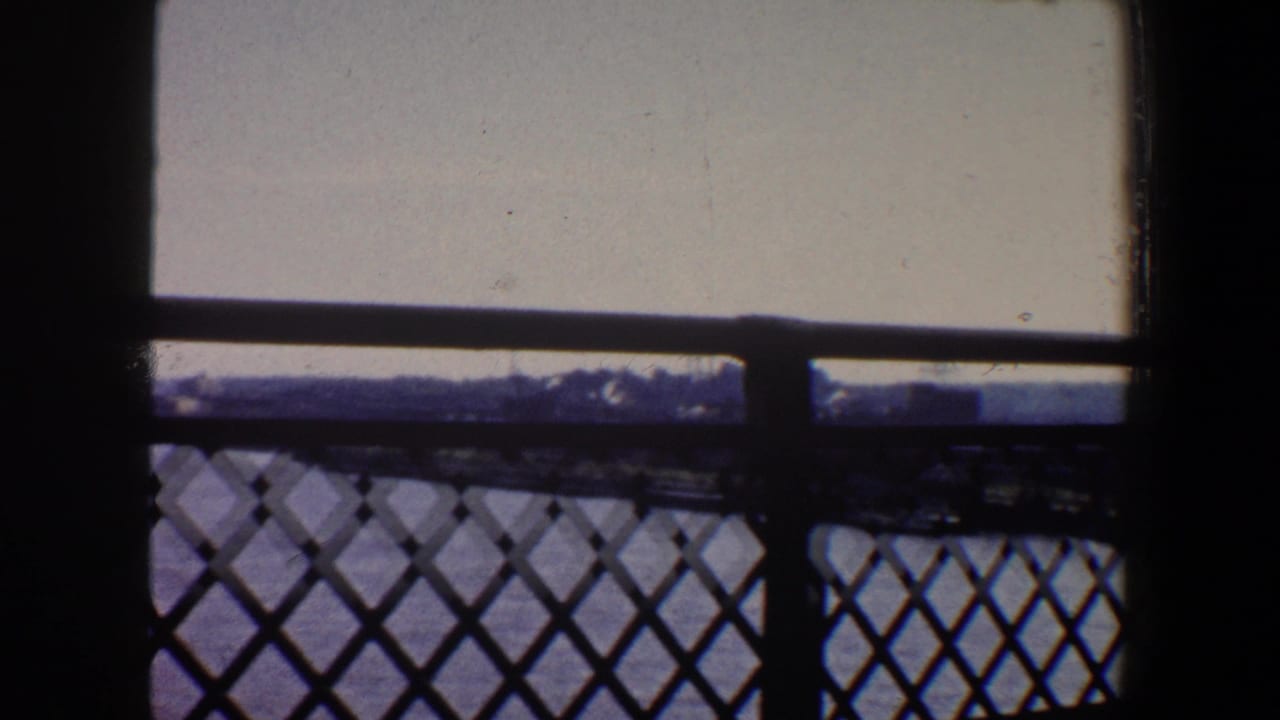 1964 Saint Louis 8mm frame looking from shadow through a diamond-pattern metal railing toward water, pale sky, and distant forms.