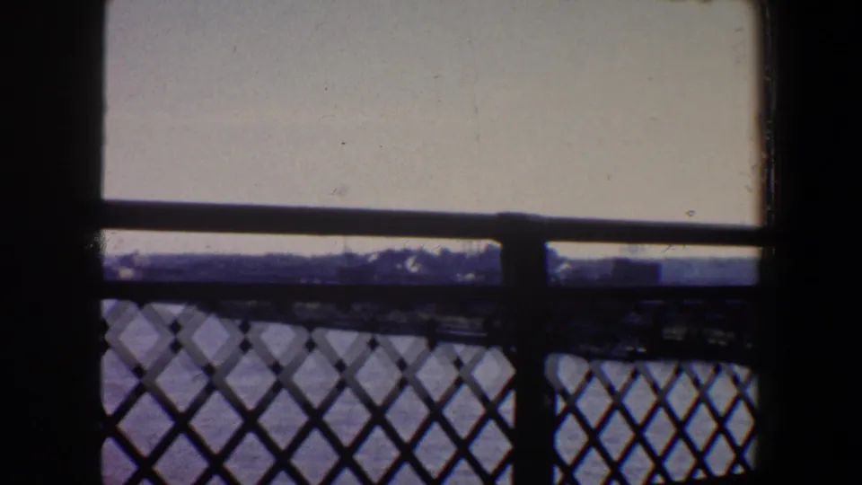 1964 Saint Louis 8mm frame looking from shadow through a diamond-pattern metal railing toward water, pale sky, and distant forms.