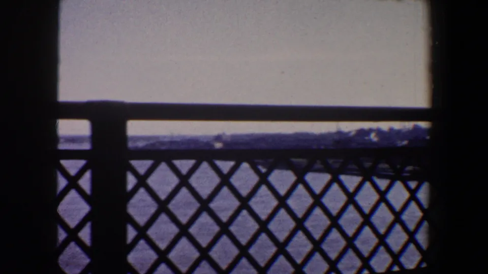 1964 Saint Louis home-movie frame with a silhouetted metal railing crossing a view of water, pale sky, and distant shoreline or structures.