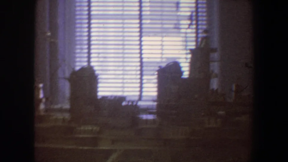 1964 Saint Louis 8mm frame of a dark interior with bright horizontal blinds and indistinct silhouetted objects in the foreground.