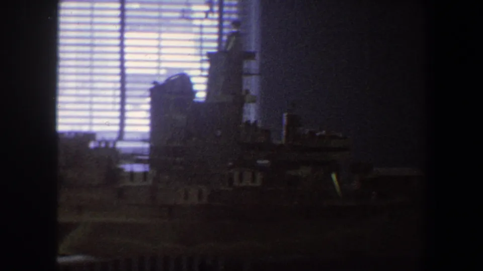 1964 Saint Louis home-movie frame showing a dim room, window blinds, and a shadowed model naval ship on a table.