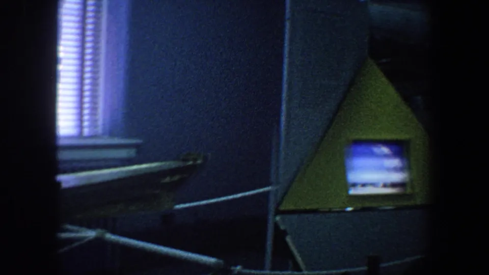 1964 Saint Louis home-movie frame with a dim model ship foreground and a triangular display-like object near a window with blinds.