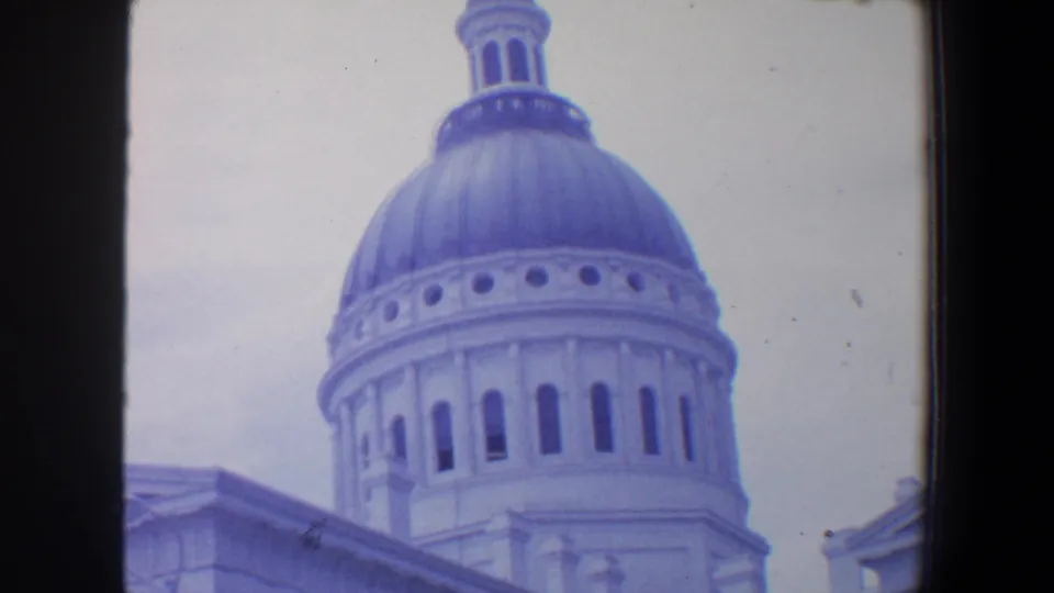 1964 8mm frame of a close exterior view of a domed classical building with roofline and architectural details visible below.