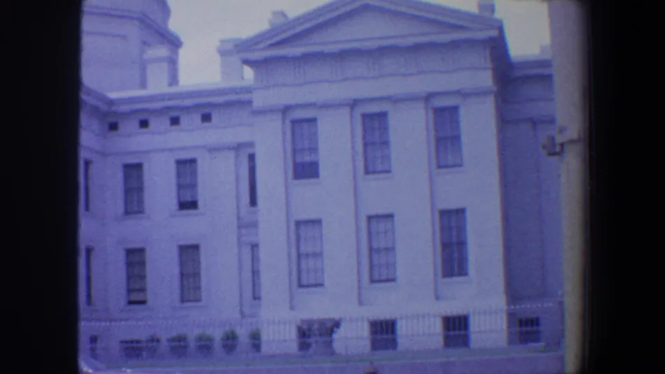 1964 Saint Louis 8mm frame showing the lower facade, tall windows, iron fence, and street-level grounds of a classical building.