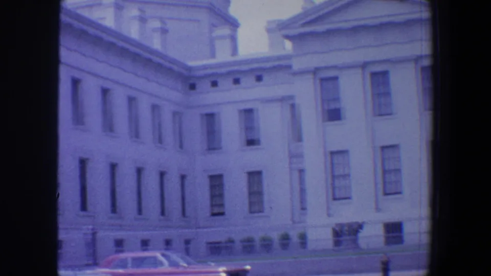 1964 Saint Louis home-movie frame with a white domed classical building and a red period car passing near the street edge.