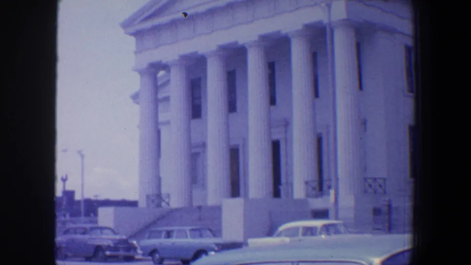 Saint Louis 1964 exterior with a white columned building, lamppost, and several mid-century cars along the street.