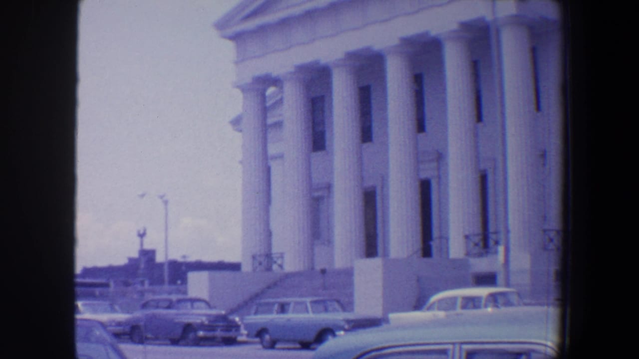 Columned white building facade with parked 1960s cars under a cloudy sky.