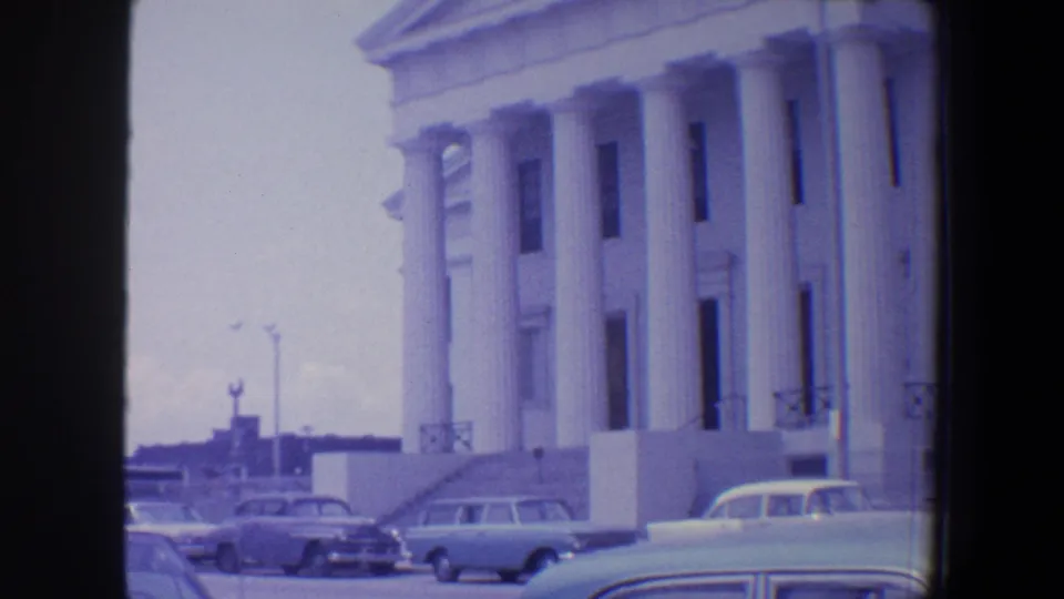 1964 Saint Louis home-movie frame showing a white columned civic building, parked period cars, and cloudy daylight.
