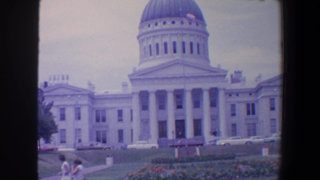 Domed columned civic building with lawns, parked cars, flower beds, and two pedestrians in front.