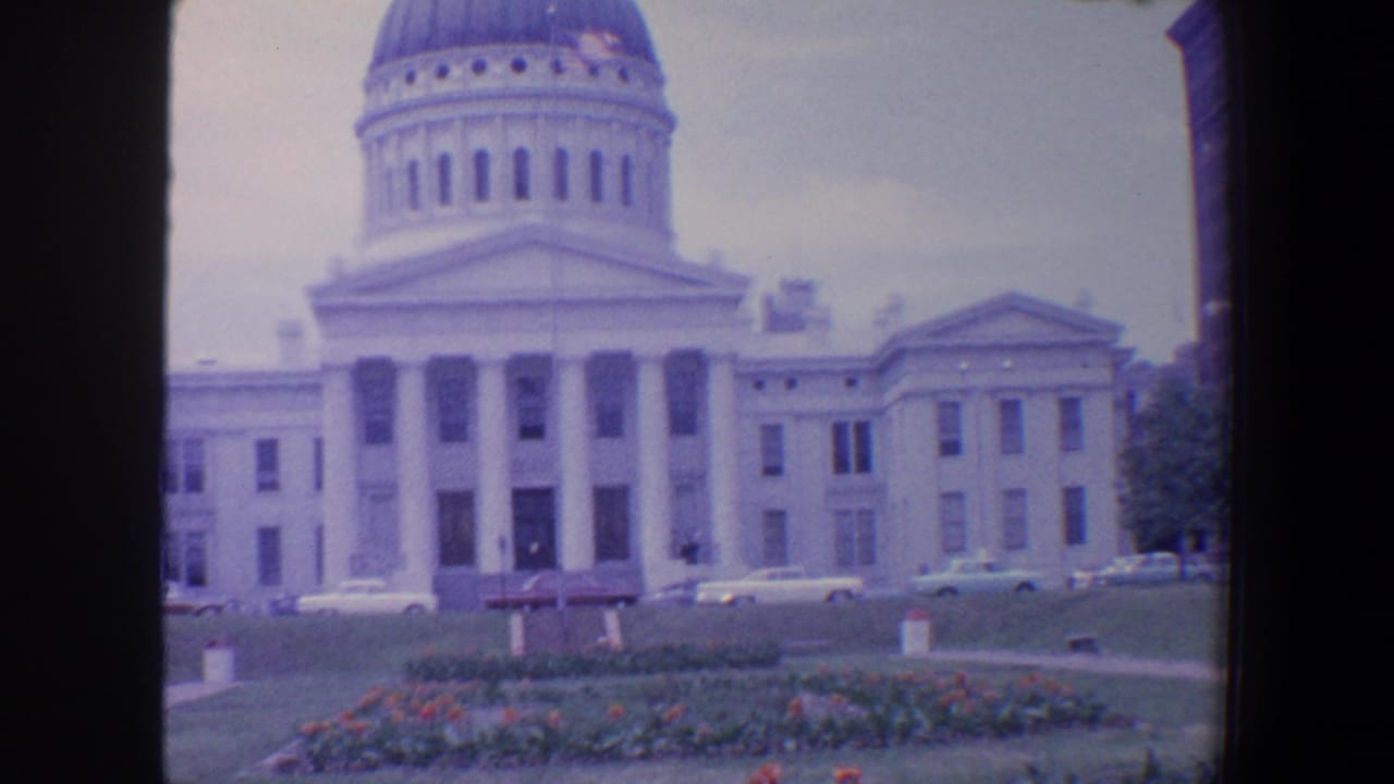 Domed columned building seen across a lawn with cars and landscaped beds in front.