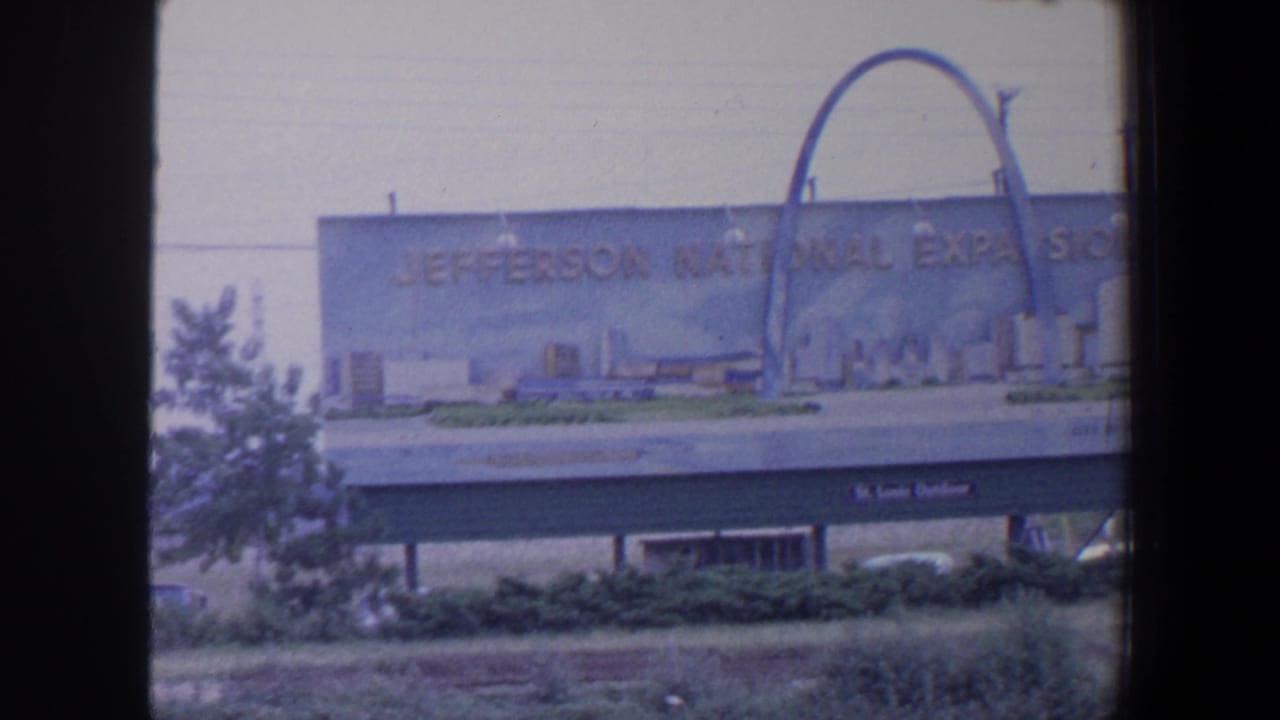 Billboard for the Jefferson National Expansion Memorial beside a road, with Gateway Arch artwork and trees nearby.