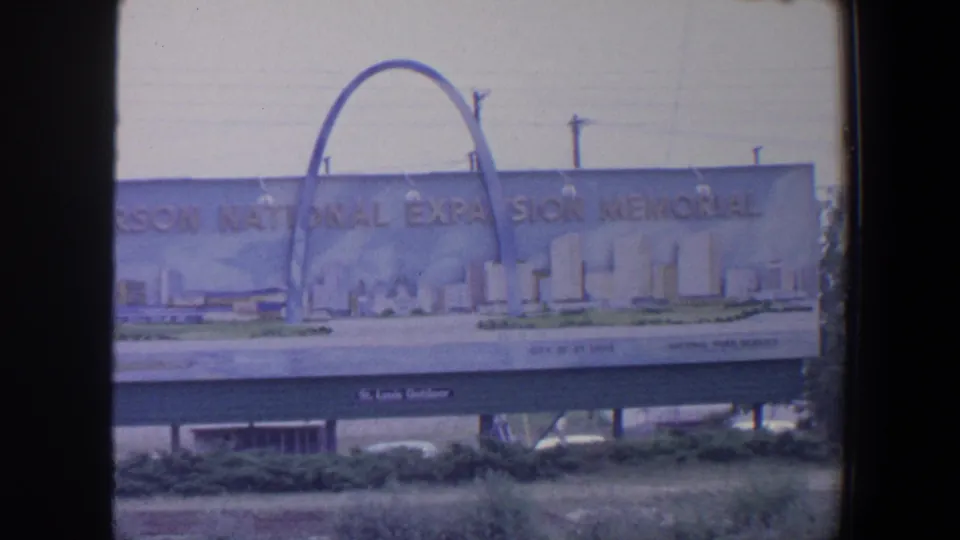 1964 St. Louis 8mm frame showing a closer roadside view of the Jefferson National Expansion Memorial billboard and its Gateway Arch illustration.