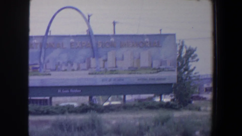 1964 St. Louis home-movie frame with the memorial billboard partly shifted in view, showing arch artwork, supports, grass, and a roadside tree.