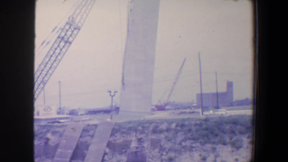1964 Saint Louis home-movie frame showing a crane beside a tall pale Gateway Arch construction segment under cloudy sky.
