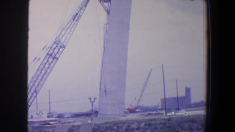 Saint Louis 1964 frame with a pale Gateway Arch segment, crane boom, smaller cranes, and open construction ground.