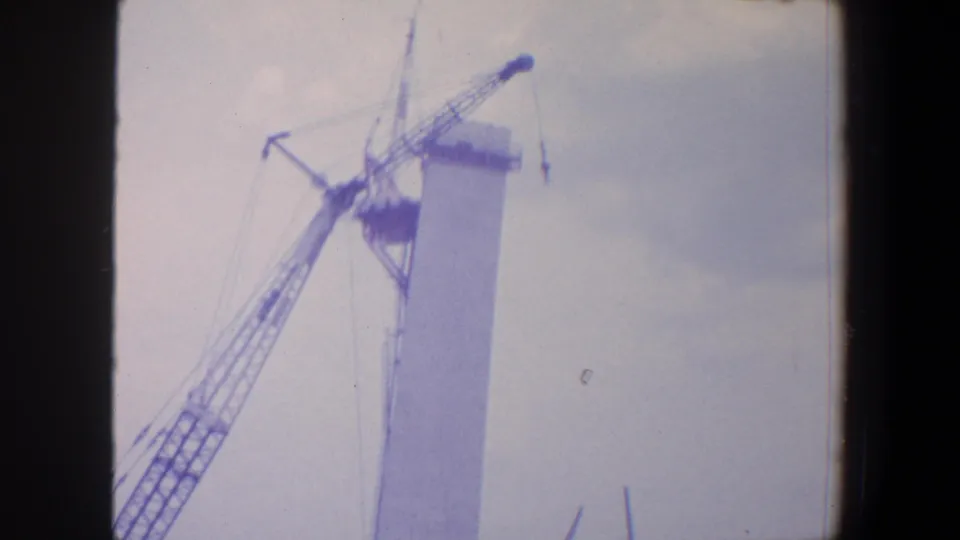 Saint Louis 1964 home-movie frame showing crane machinery beside a Gateway Arch construction segment, with cloudy sky and soft film color.
