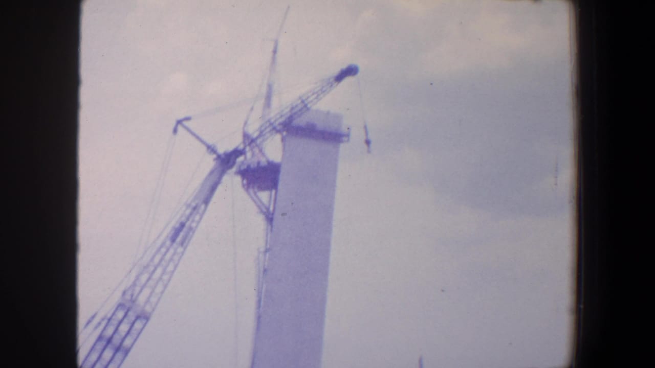 1964 Saint Louis home-movie frame showing a crane beside a tall industrial tower under construction against a washed pale sky.
