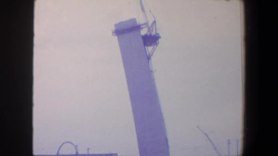 Handheld 1964 Saint Louis frame of a leaning view toward a tall construction tower, with part of the Gateway Arch visible near the lower edge.