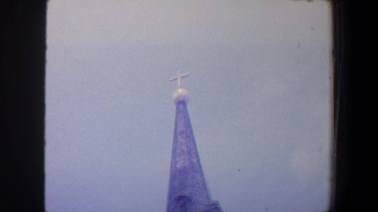 Soft low-angle view of a church steeple top and cross against pale sky.