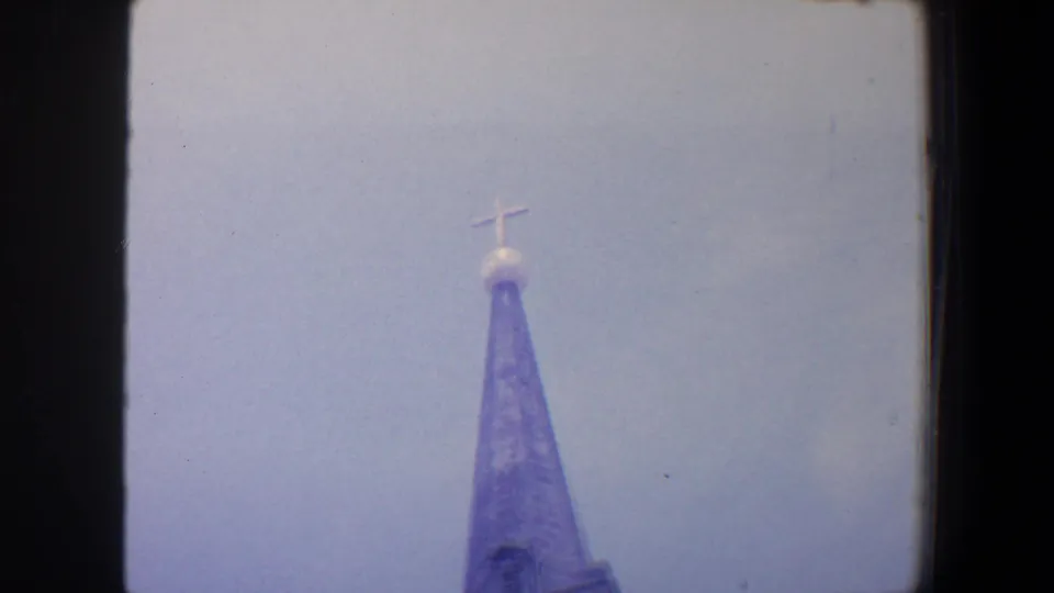 1964 Saint Louis 8mm frame showing the upper portion of a grey church steeple with a cross against a pale sky.