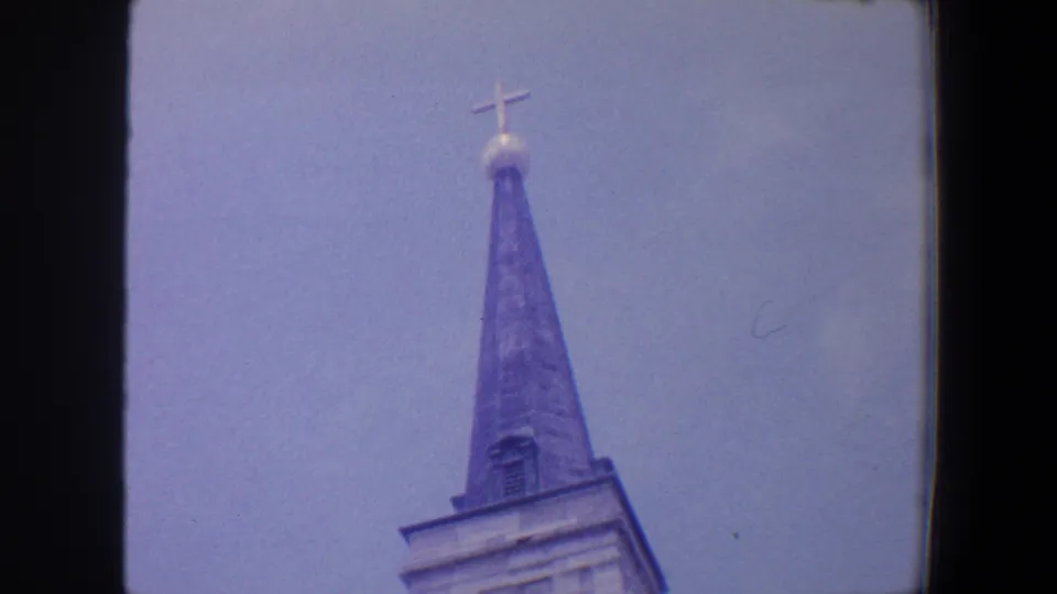 1964 Saint Louis home-movie frame with a grey church steeple, cross, orb, and upper tower visible against blue sky.