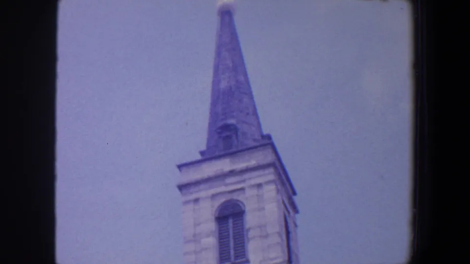 1964 Saint Louis 8mm still showing a grey church tower with an arched louvered window and steeple topped by a cross.