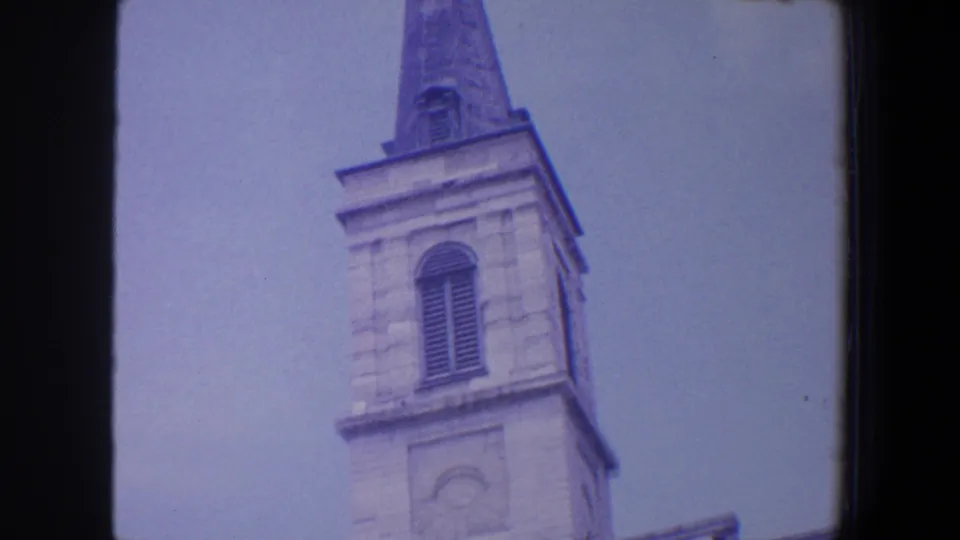 1964 Saint Louis frame showing the church tower, arched window, tall steeple, orb, and cross in a low-angle composition.