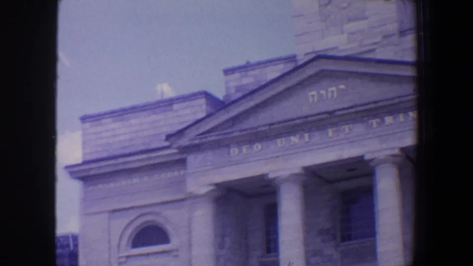 1964 Saint Louis frame showing a pale stone religious building facade with tower, columned portico, arched window, Hebrew letters, and Latin motto.