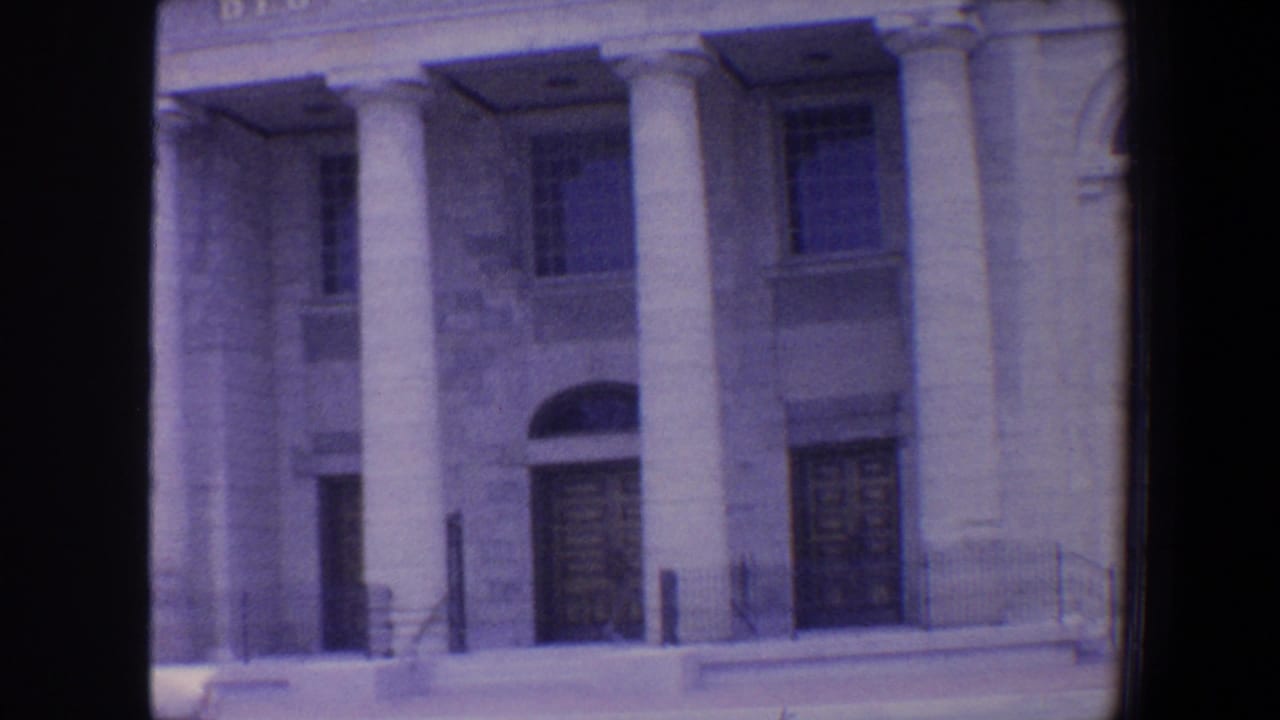 Stone building facade with columns, arched doorway, windows, steps, and street curb.