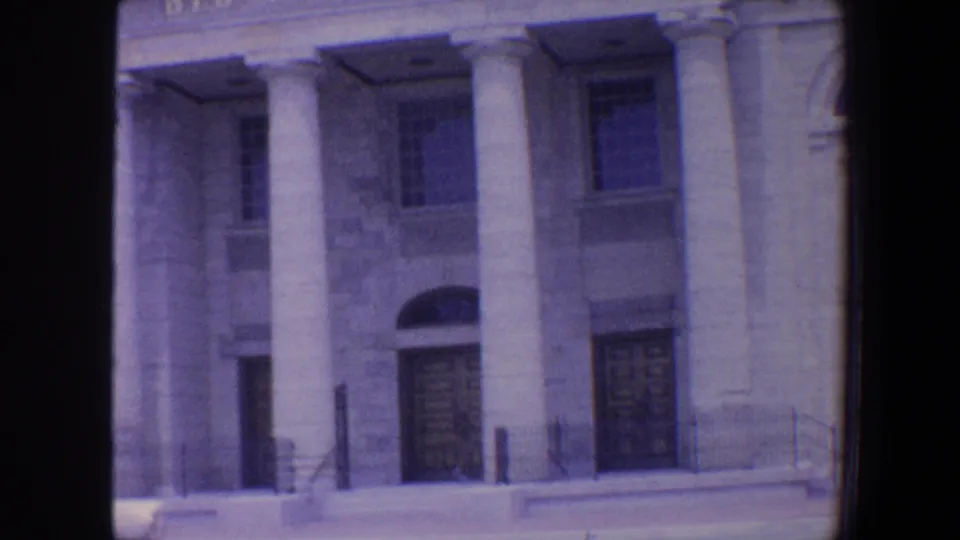 1964 Saint Louis home-movie frame showing a formal stone facade with columns, arched wooden door, tall windows, steps, and curb.