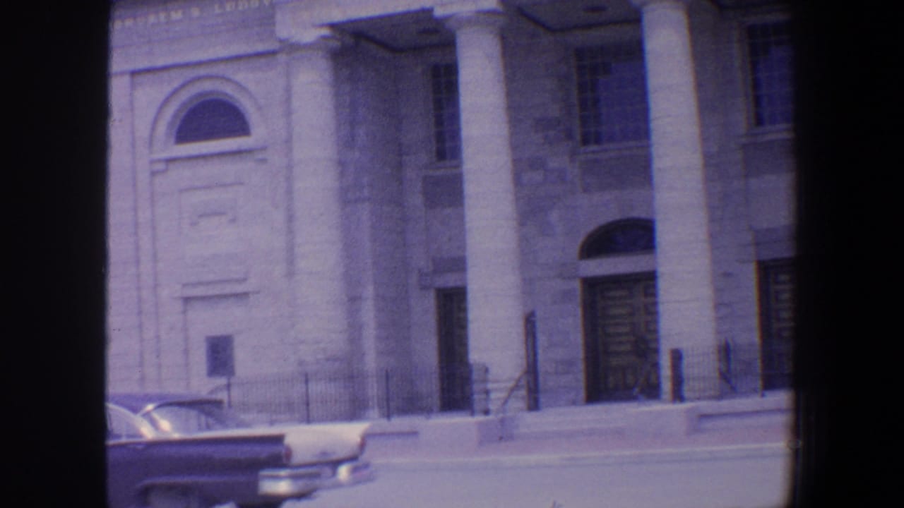 Classical stone facade with arched entry, columns, windows, railings, and a partial parked car.