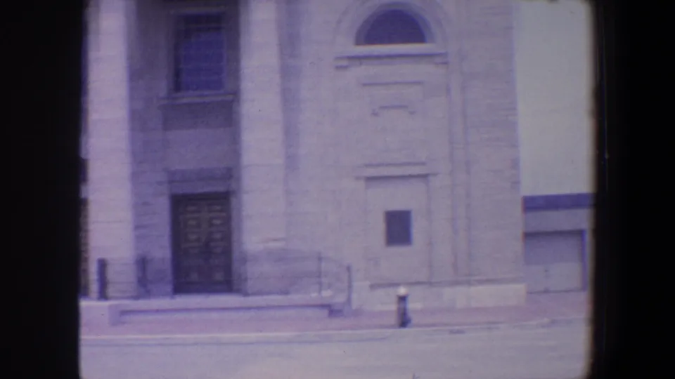 1964 Saint Louis frame showing a stone building section with an arched upper detail, doorway, plaque, steps, and sidewalk curb.