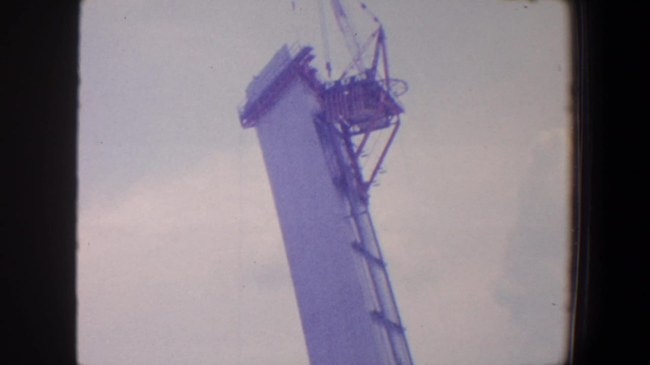 Upper section of a tapered construction tower with crane lines and dark film border.