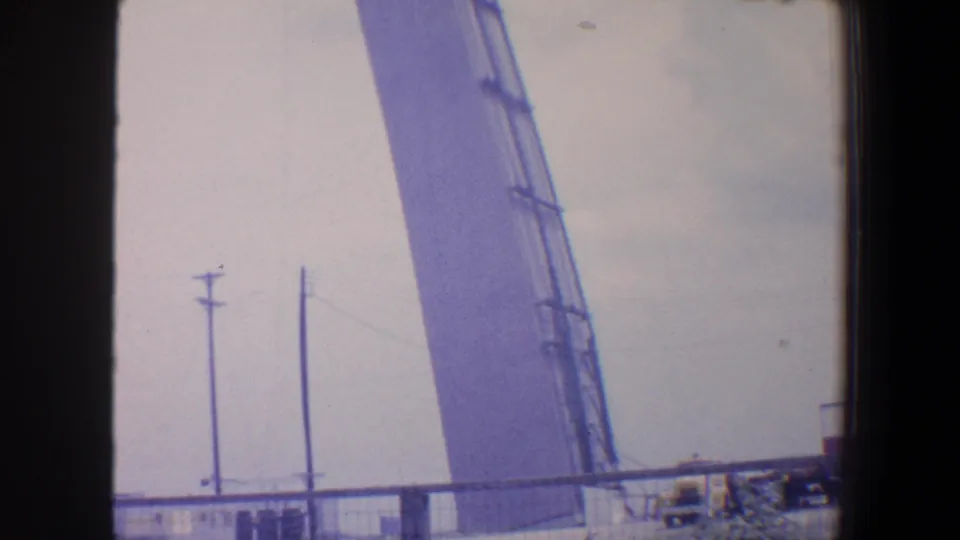 1964 Saint Louis home-movie frame showing a large angled industrial structure, utility poles, fencing, and pale sky through dark window framing.