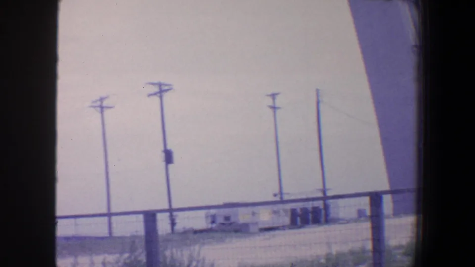 1964 Saint Louis frame showing fencing, utility poles, low industrial structures, and the side of a large angled support in soft 8mm color.