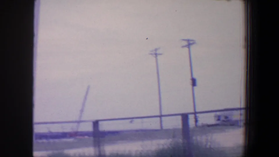 1964 Saint Louis home-movie frame with utility poles, a fence line, low structures, and a distant angled industrial element under a pale sky.