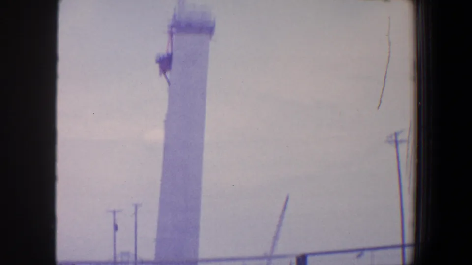 1964 Saint Louis home-movie still focused on the top of a white construction column with crane equipment visible.