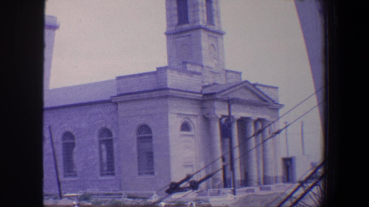 1964 Saint Louis home-movie frame showing a stone church exterior with a bell tower, arched windows, columned entrance, and scaffolding in front.